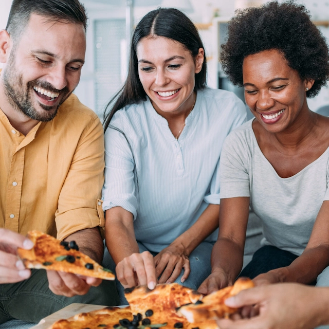 Shot of multiracial group of people sitting on the sofa in the living room and eating pizza. Two young couples having fun time and eating pizza at home.